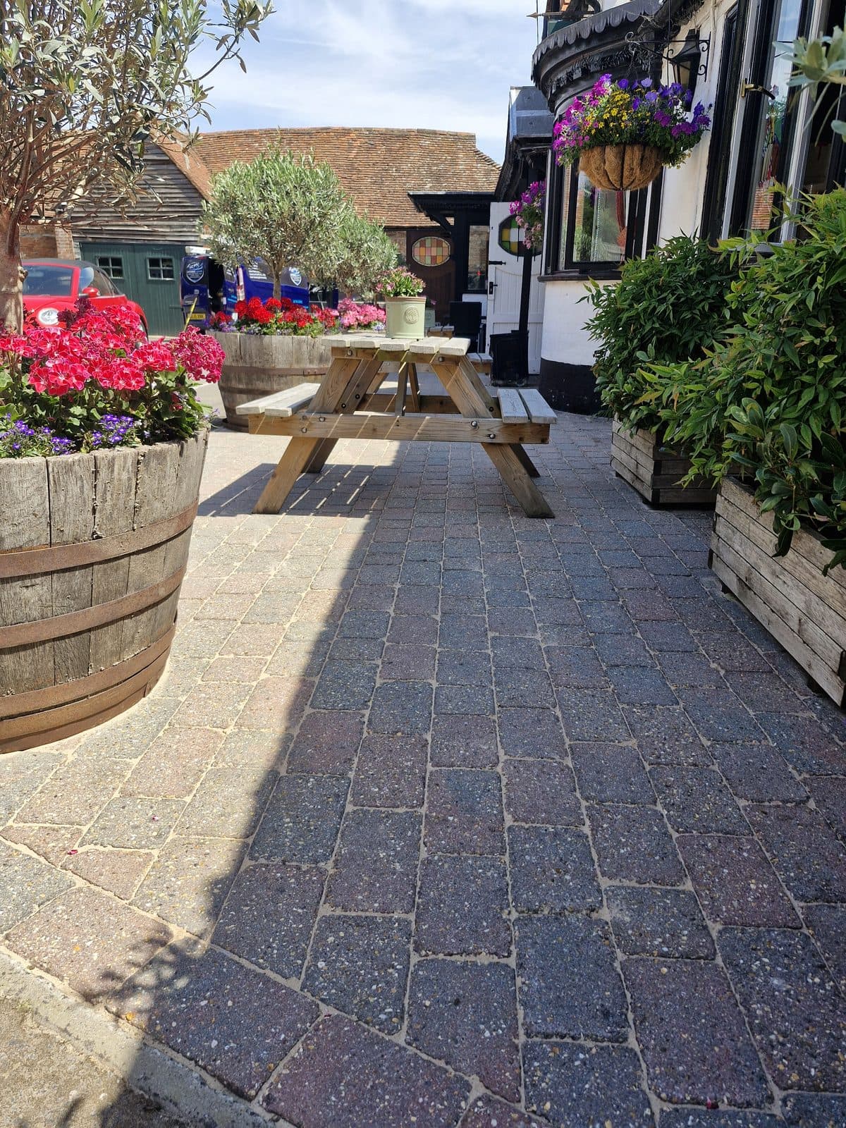 A pub courtyard of block paving, with picnic benches and barrels of red and pink flowers.